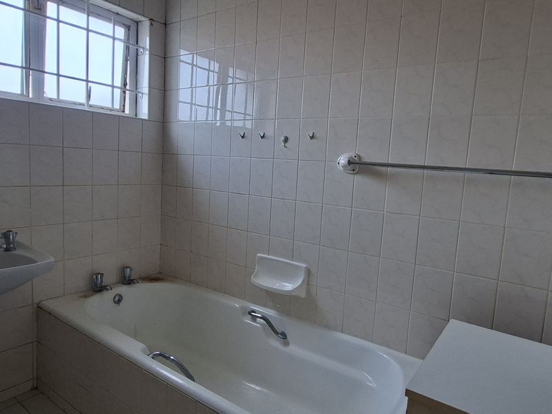 A dated bathroom featuring a pedestal basin, built-in bathtub, and full-height wall tiling, requiring cosmetic updates to modernize the space.