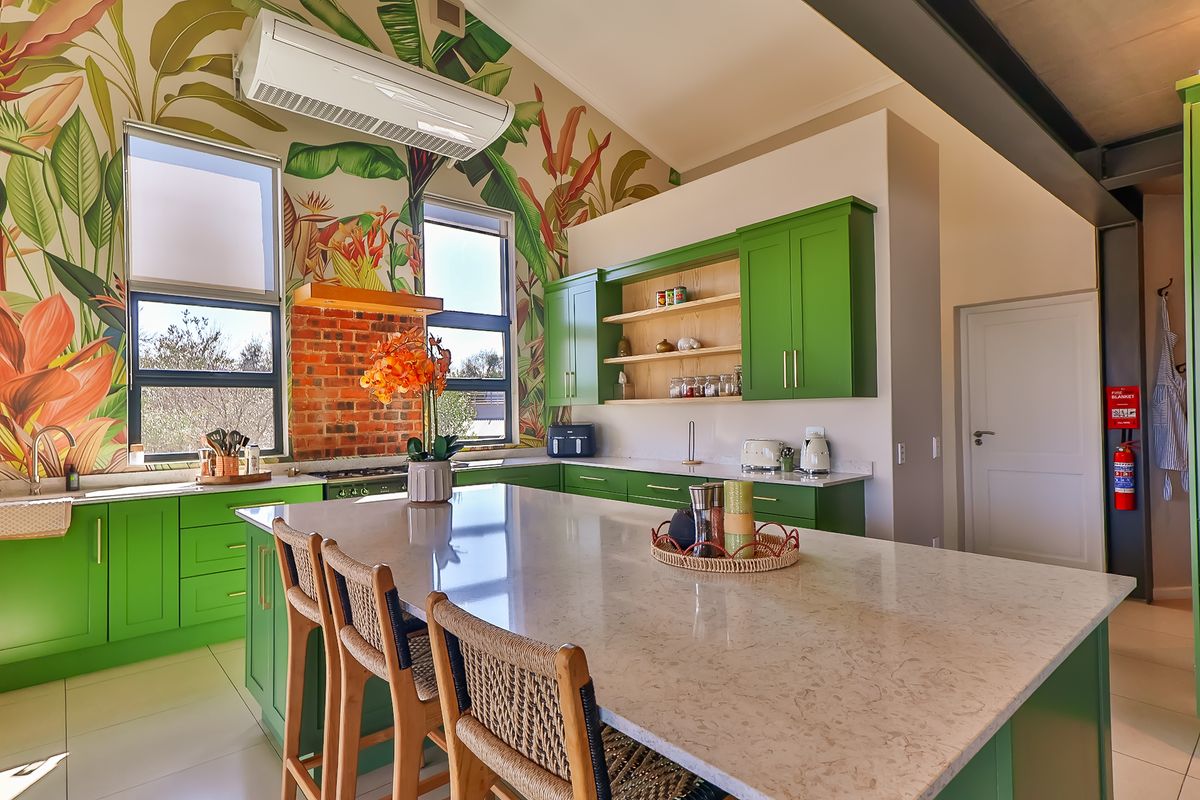 A vibrant, modern kitchen featuring green cabinetry, a large stone-topped island, exposed brick accents, and striking botanical wallpaper under a vaulted ceiling.