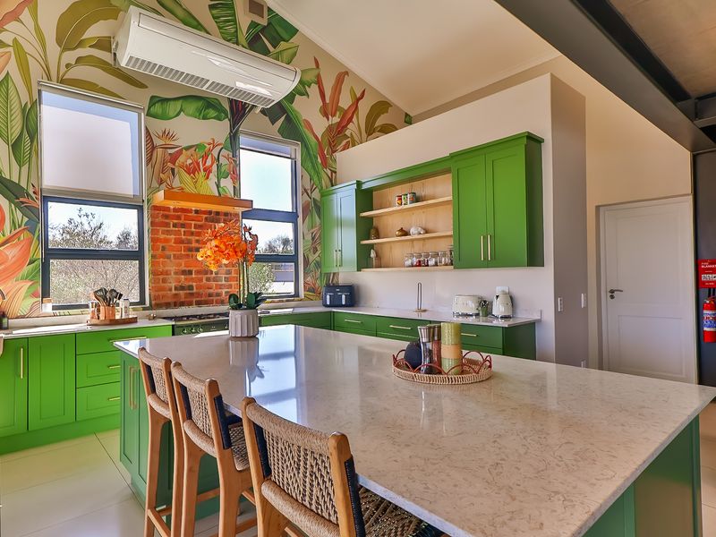 A vibrant, modern kitchen featuring green cabinetry, a large stone-topped island, exposed brick accents, and striking botanical wallpaper under a vaulted ceiling.