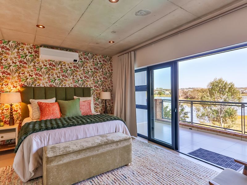 Spacious bedroom featuring an exposed concrete ceiling, split-system air conditioning, and large sliding glass doors opening onto a private balcony with scenic water views.