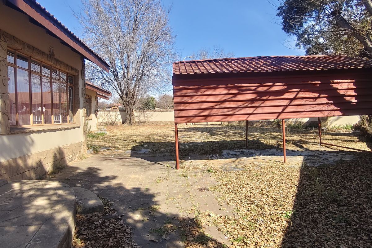 Rear exterior view featuring a paved patio area, a metal carport structure, and a spacious garden with boundary walling.