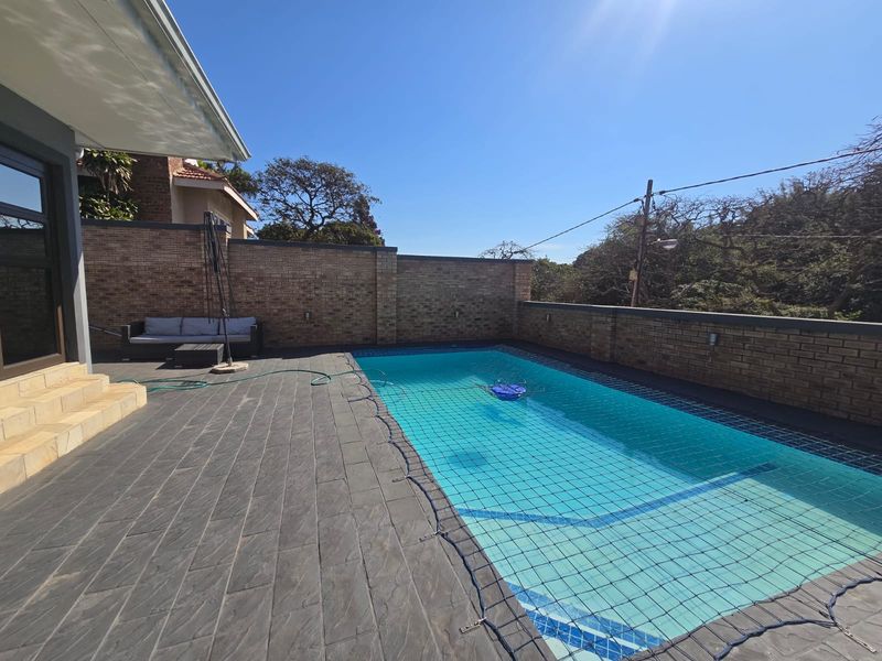 A well-maintained outdoor pool area featuring a tiled deck, brick boundary walls, and a safety net over the swimming pool.