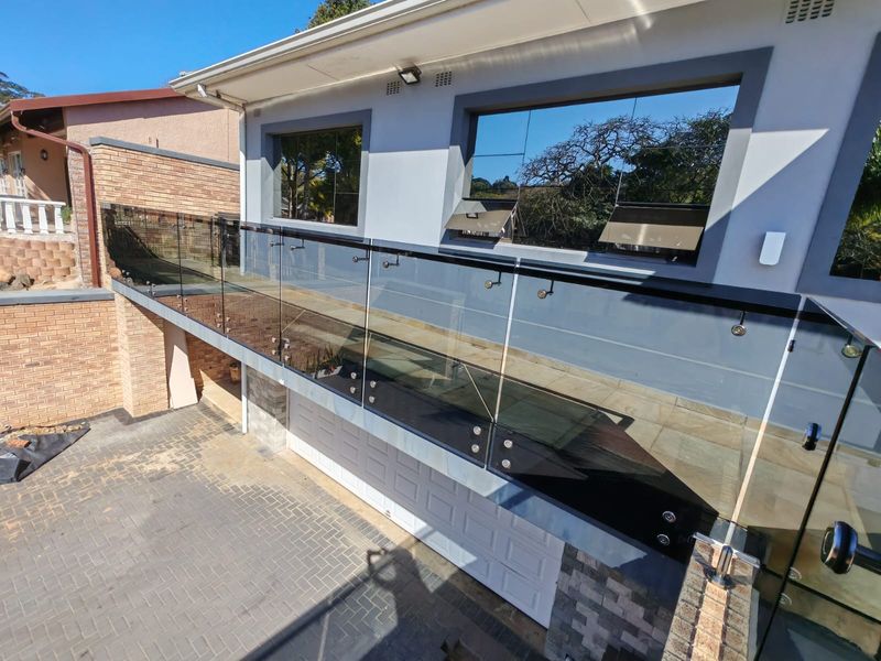 A modern elevated balcony featuring a sleek frameless glass balustrade, overlooking a paved driveway and garage area.