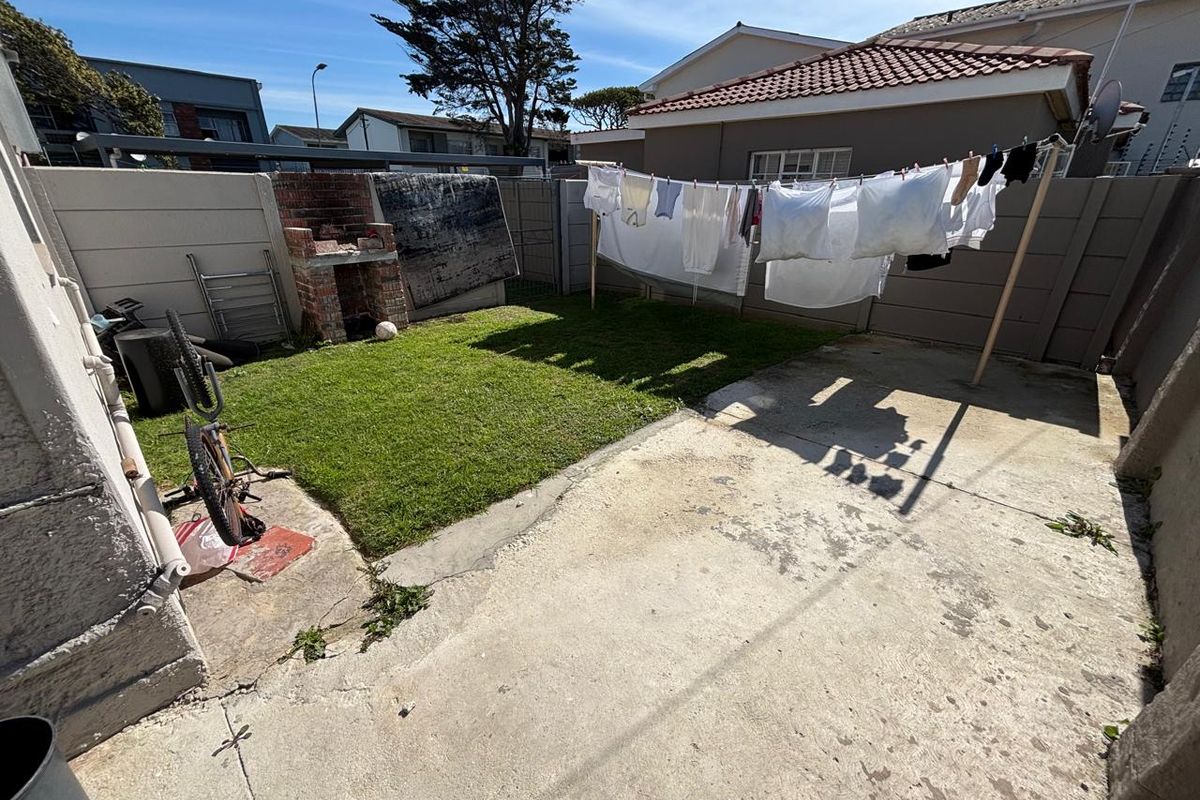 A rear garden area featuring a concrete patio, a small lawn, and a built-in brick braai, enclosed by precast concrete boundary walls.