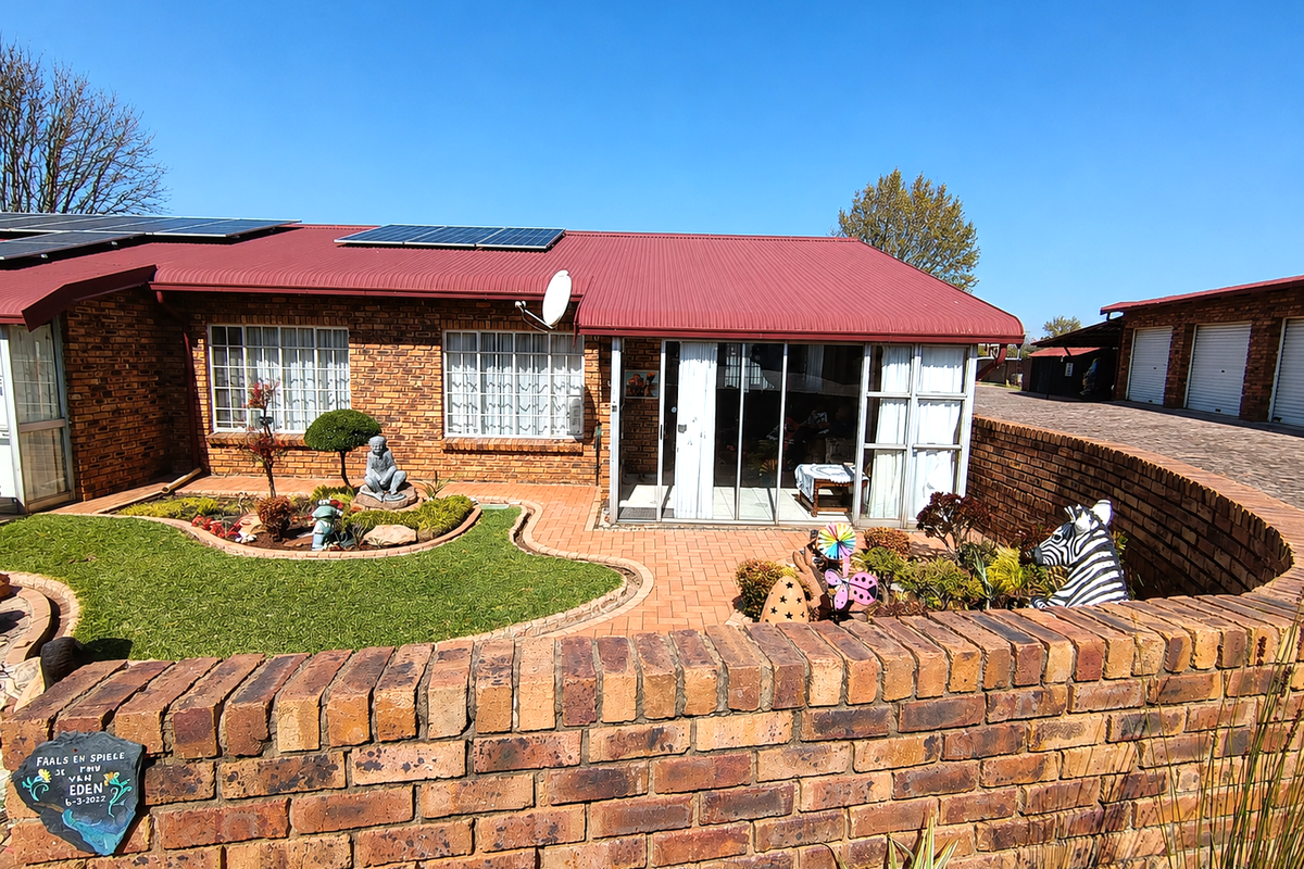 A well-maintained face brick apartment exterior featuring a red corrugated metal roof, solar panels, a paved patio area, and a private garden.