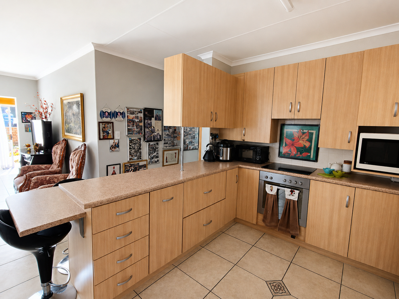 Open-plan kitchen featuring light wood-finish cabinetry, laminate countertops, a breakfast bar, and integrated oven and hob, flowing into the adjacent living area.