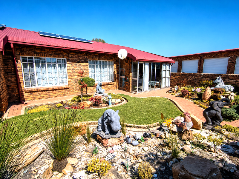 Rear view of the property featuring a brick facade, corrugated metal roof with solar panels, and a landscaped garden with a paved walkway.