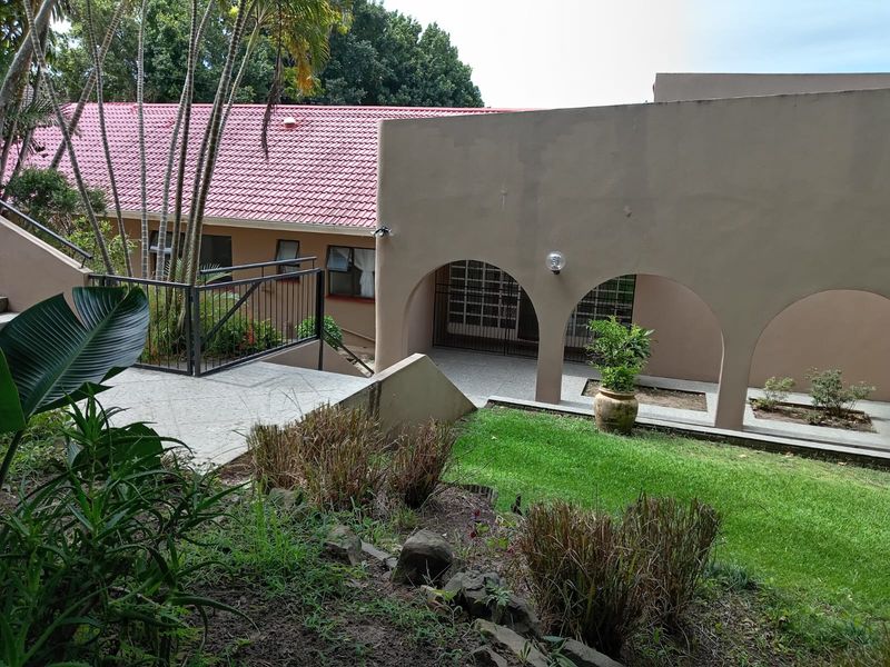 Rear exterior view of the property featuring a covered arched walkway, tiled roof, and landscaped garden area with retaining walls.