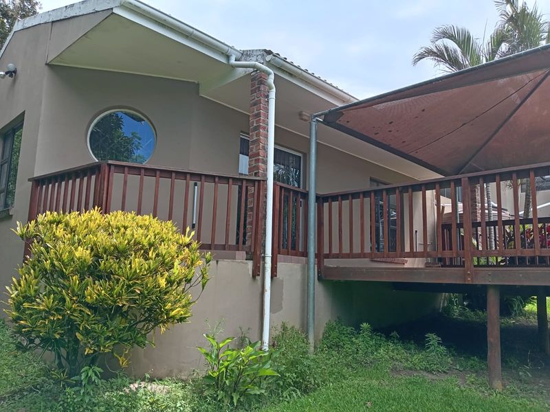 Rear exterior view featuring a raised wooden deck with timber balustrades, a circular window, and a shade sail structure over the outdoor area.