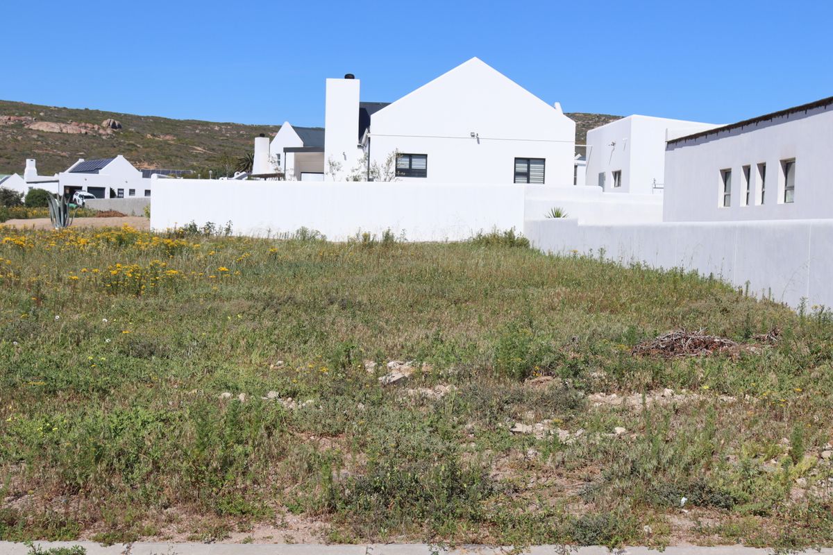 A vacant residential plot featuring natural vegetation, enclosed by a white boundary wall with neighboring properties visible in the background.