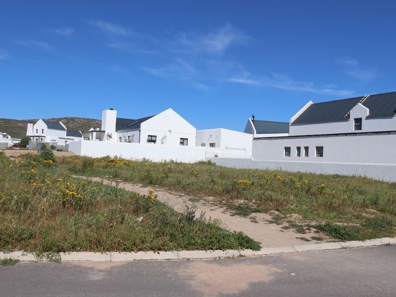 A vacant residential plot situated in a developed estate, featuring natural vegetation and bordering established white-walled homes.