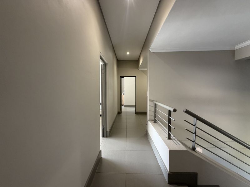 A clean, modern hallway featuring neutral tiled flooring, recessed lighting, and a contemporary metal balustrade overlooking the lower level.