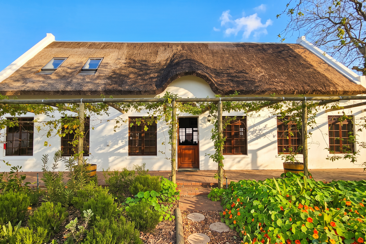 Charming Cape Dutch style home featuring a traditional thatched roof, timber pergola, and a well-maintained brick patio overlooking a lush garden.