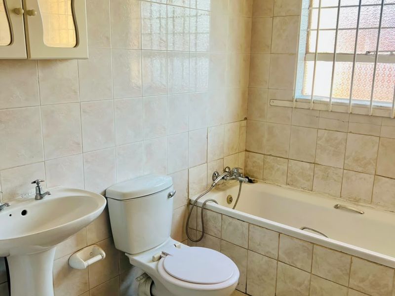 A dated bathroom featuring a pedestal basin, toilet, and bathtub with full-height wall tiling and security bars on the window.