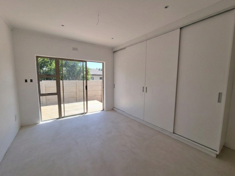 A bright, empty bedroom featuring built-in sliding door wardrobes, concrete flooring, and a sliding glass door providing direct access to an outdoor deck.
