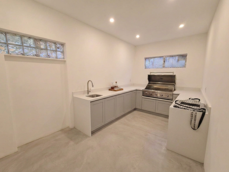 A modern, minimalist kitchen or outdoor prep area featuring sleek shaker cabinetry, quartz countertops, a built-in grill, and polished concrete floors.