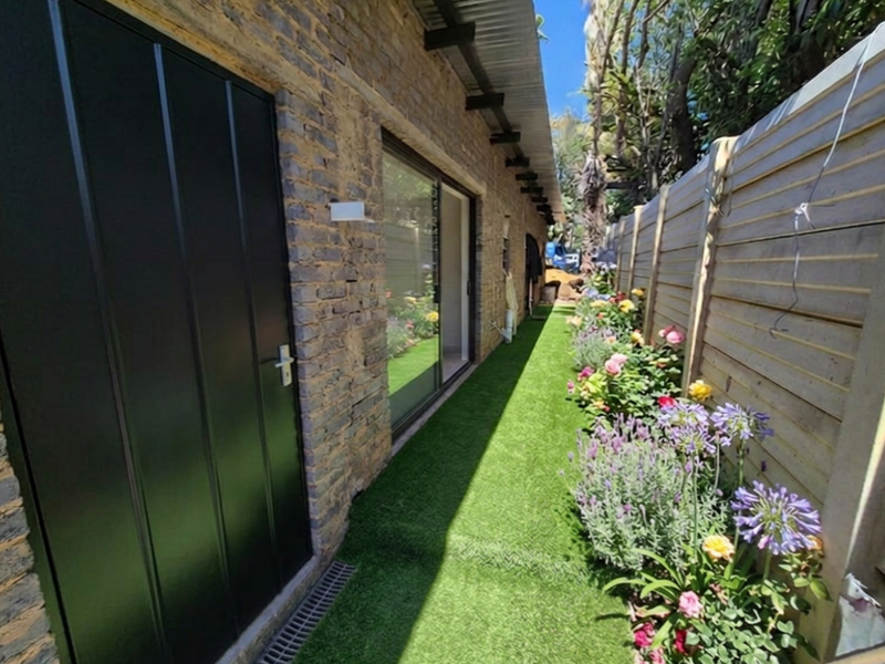 A well-maintained rear exterior view of the cottage featuring exposed brickwork, a sliding glass door, artificial turf, and a landscaped garden border.