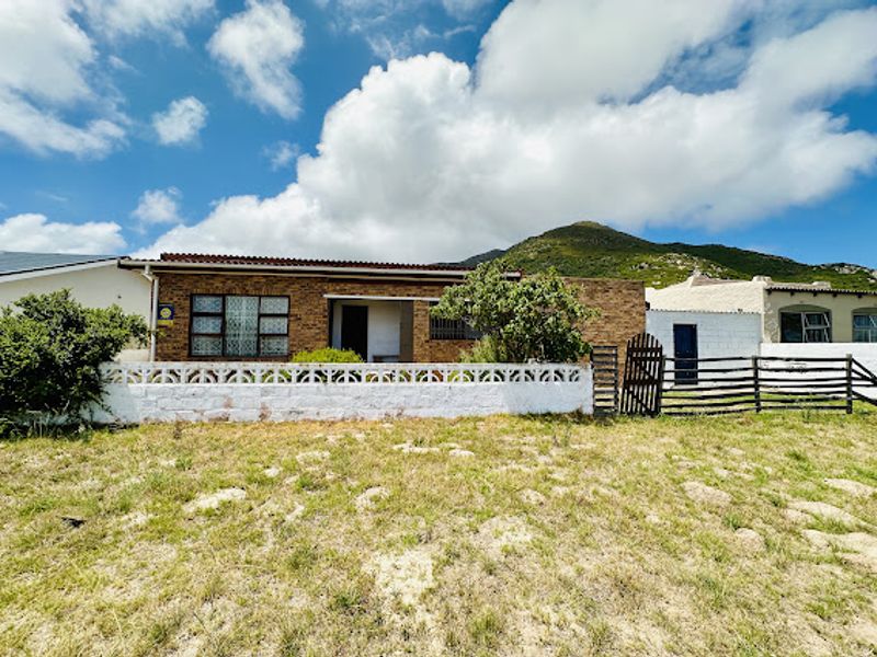 Front elevation of a single-story brick house featuring security bars, a low boundary wall, and a timber gate, set against a mountain backdrop.