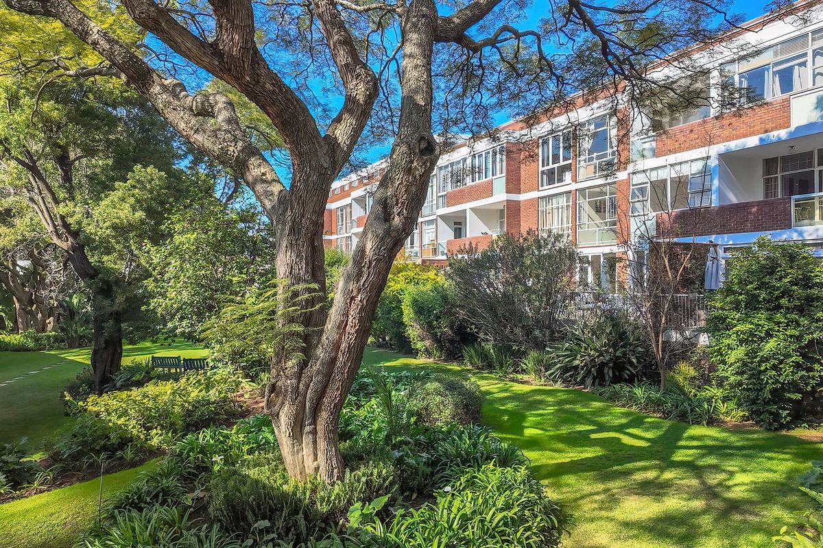 A view of the apartment building's rear facade featuring brick construction, private balconies, and well-maintained communal garden areas with mature trees.