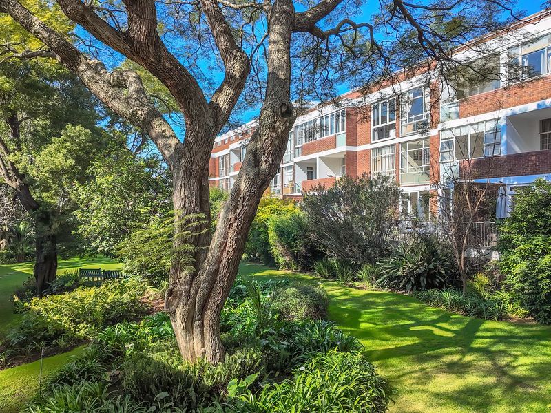 A view of the apartment building's rear facade featuring brick construction, private balconies, and well-maintained communal garden areas with mature trees.