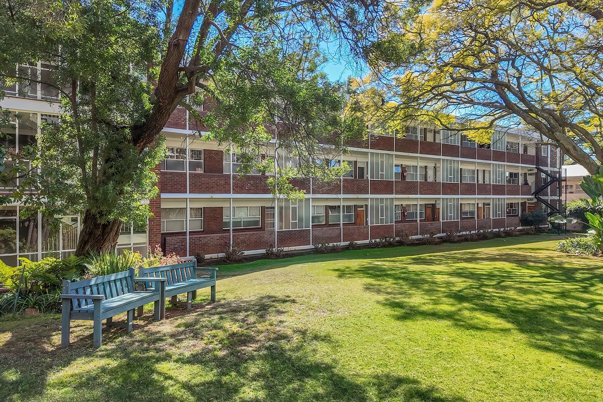 A view of the rear apartment building facade featuring brick construction, large windows, and a well-maintained communal garden area with mature trees.