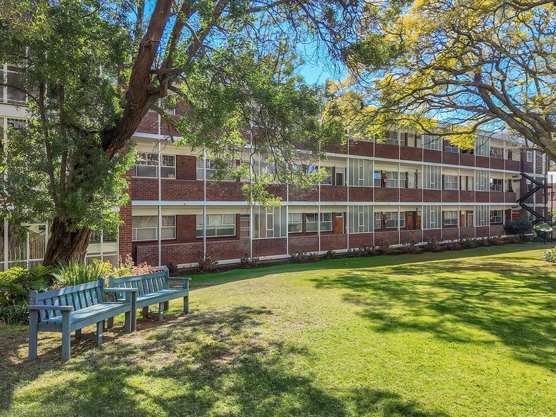 A view of the rear apartment building facade featuring brick construction, large windows, and a well-maintained communal garden area with mature trees.