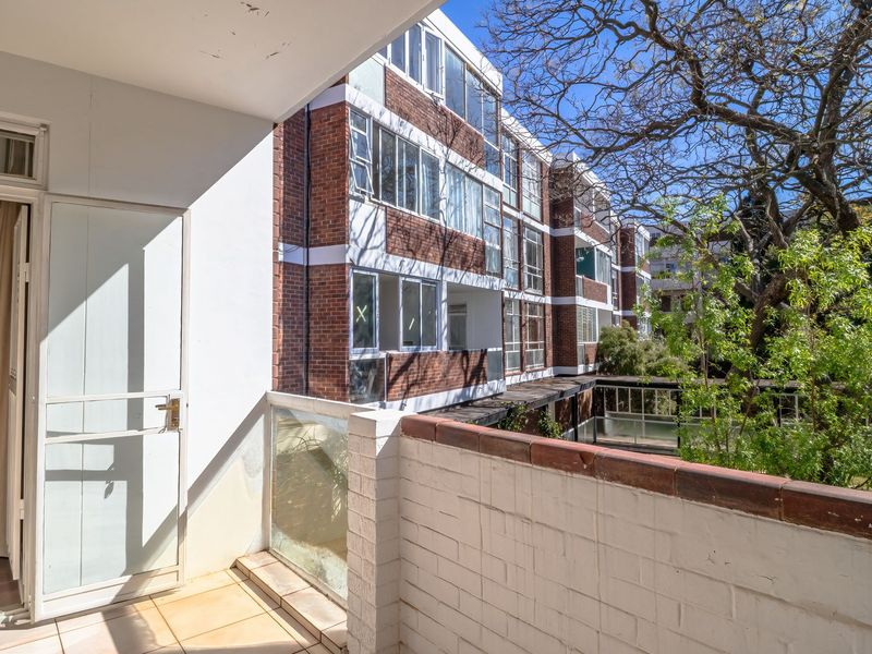 A private balcony featuring tiled flooring, a brick balustrade with a glass insert, and a security gate, overlooking the apartment complex grounds.