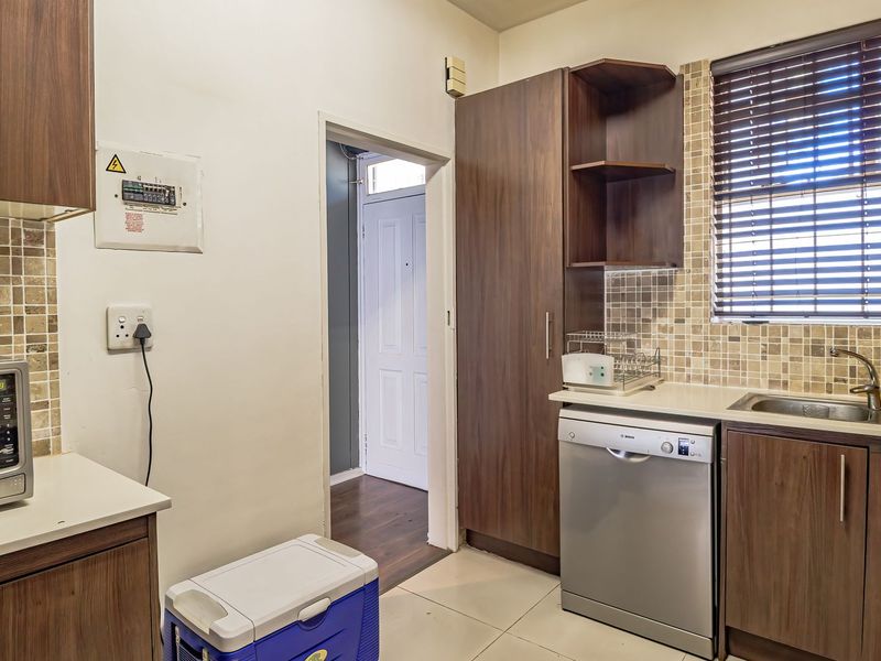 A functional kitchen featuring dark wood cabinetry, mosaic tile backsplash, stainless steel sink, and integrated dishwasher, with a view into the adjacent hallway.
