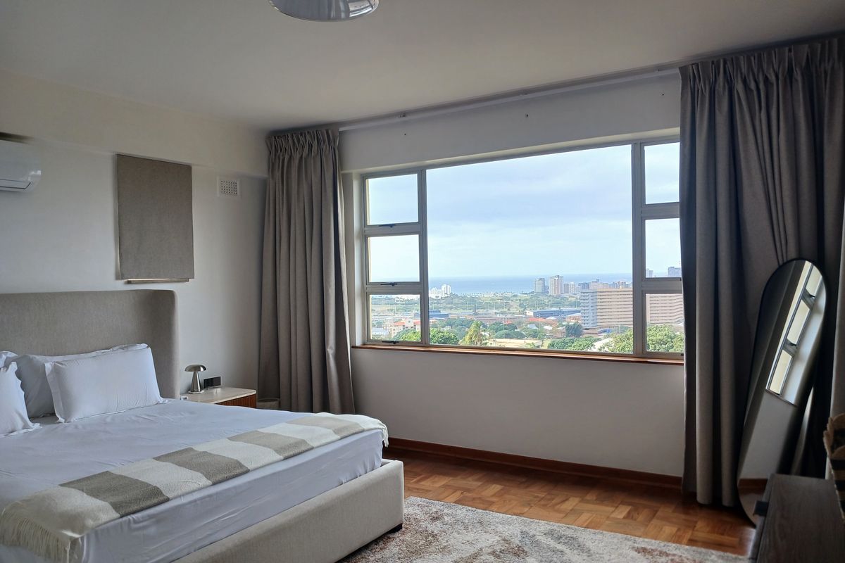 Spacious bedroom featuring warm parquet flooring, a wall-mounted air conditioner, and a large window offering expansive coastal and city views.