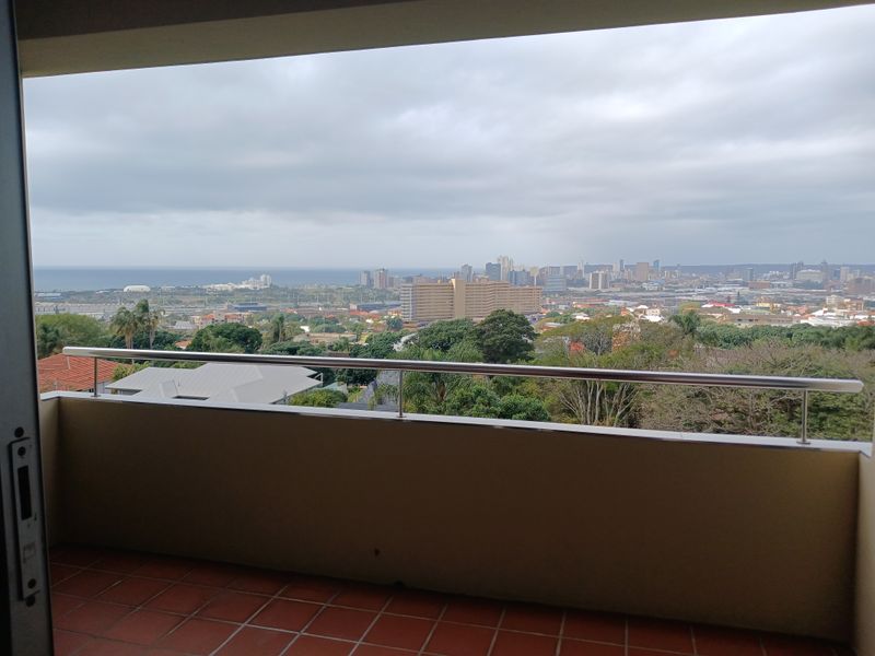 A covered balcony featuring tiled flooring and a stainless steel balustrade, offering an expansive elevated view of the city skyline and surrounding landscape.