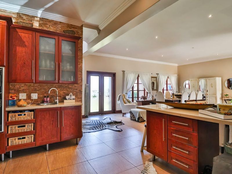 Spacious open-plan kitchen and living area featuring warm-toned wooden cabinetry, stone countertops, a feature brick wall, and tiled flooring throughout.