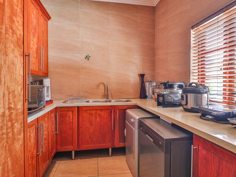 A functional scullery featuring warm-toned wooden cabinetry, stone countertops, a double basin sink, and tiled wall finishes with natural light from a window.