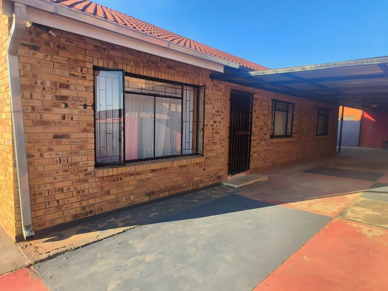 A well-maintained face brick house exterior featuring a tiled roof, secure entrance with burglar bars, and a covered carport area.