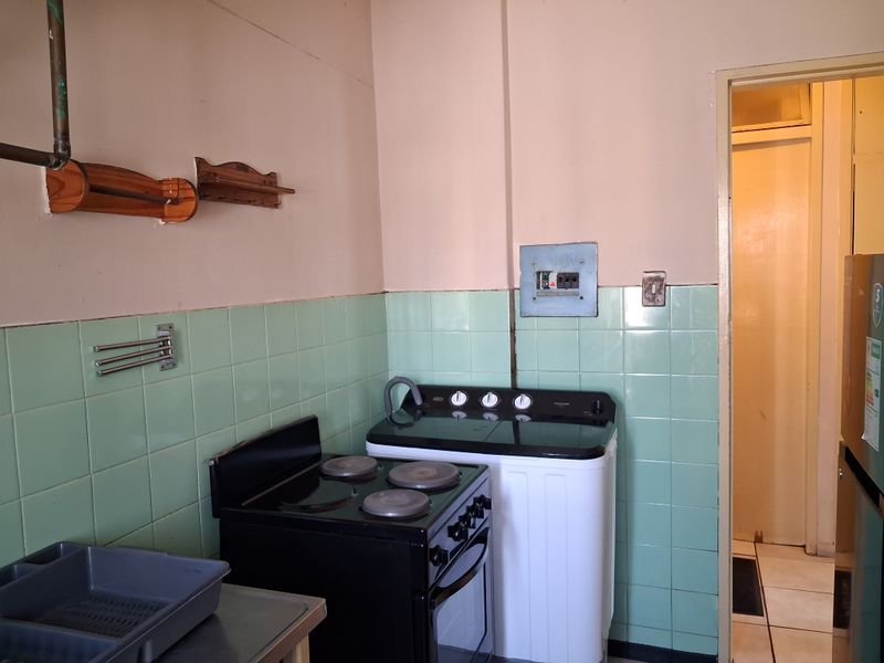 A functional kitchen area featuring a freestanding electric stove, tiled splashbacks, and an exposed electrical distribution board, requiring cosmetic updates.
