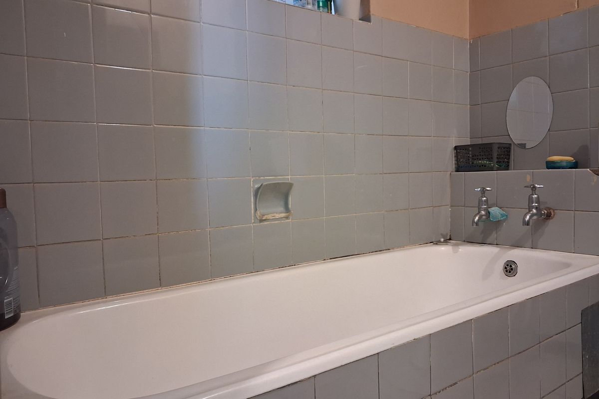 A dated bathroom featuring a standard bathtub with wall-mounted taps and grey ceramic wall tiling.