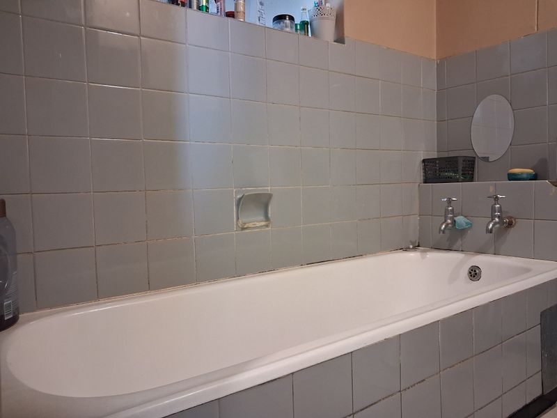 A dated bathroom featuring a standard bathtub with wall-mounted taps and grey ceramic wall tiling.