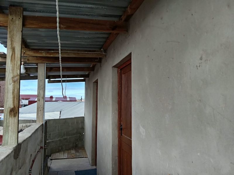 An outdoor covered walkway or balcony area featuring unfinished concrete walls, timber support beams, and a corrugated metal roof structure.