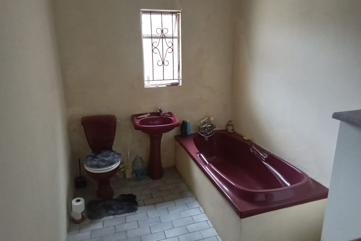 A dated bathroom featuring a burgundy suite, including a pedestal basin, built-in bathtub, and toilet, with tiled flooring and a security-barred window.