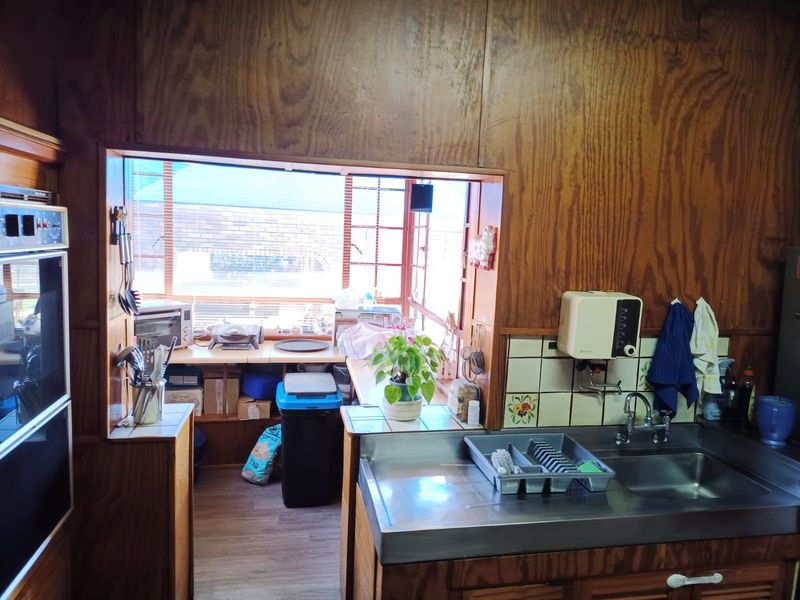 A dated kitchen featuring wood paneling, a stainless steel sink, and a built-in oven, with a pass-through window looking into an adjacent utility area.