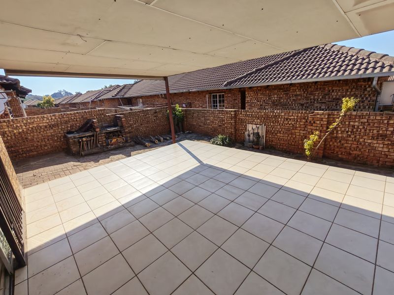 A spacious covered patio featuring tiled flooring and a built-in brick braai, enclosed by brick boundary walls in a townhouse complex.
