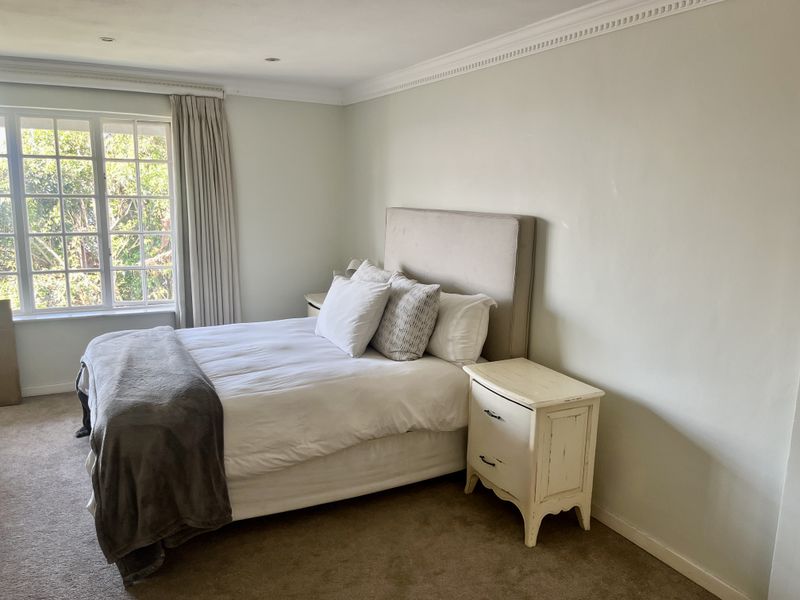 A well-lit bedroom featuring decorative crown molding, recessed ceiling lighting, and a large multi-pane window overlooking greenery.