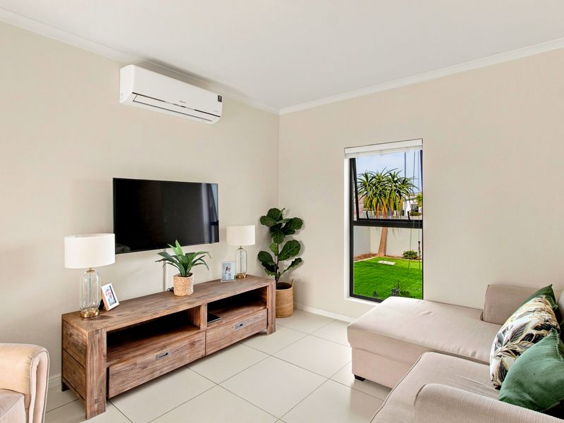 A bright living room featuring tiled flooring, a wall-mounted air conditioning unit, and a window overlooking the garden.