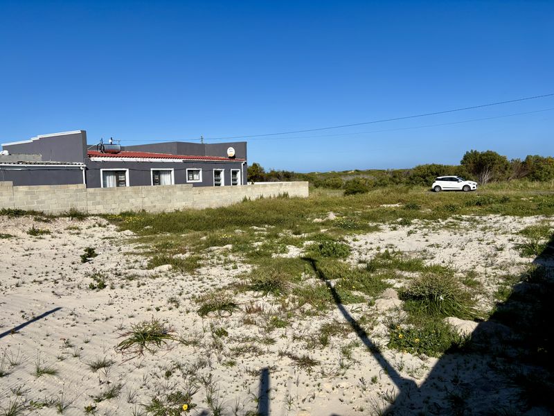 A view of a vacant residential plot featuring sandy terrain, natural vegetation, and a concrete block boundary wall adjacent to an existing dwelling.