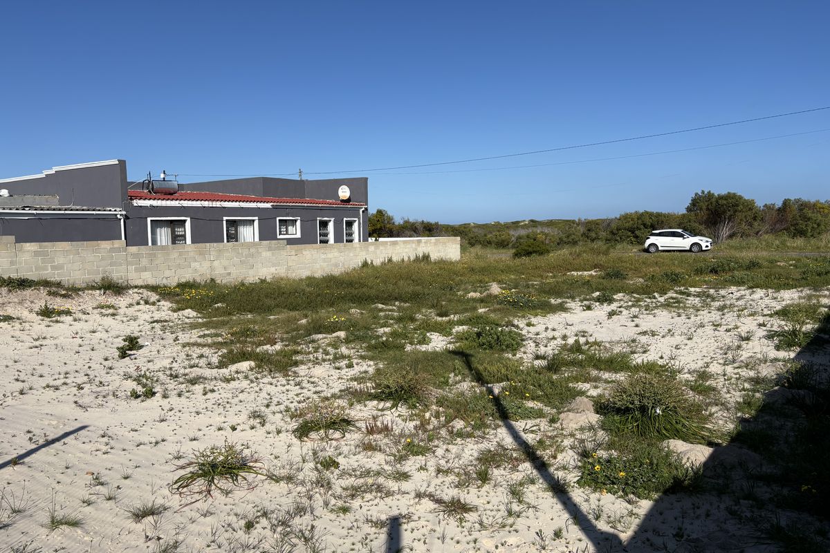 A view of a vacant residential plot featuring sandy terrain, natural vegetation, and a concrete block boundary wall adjacent to an existing structure.