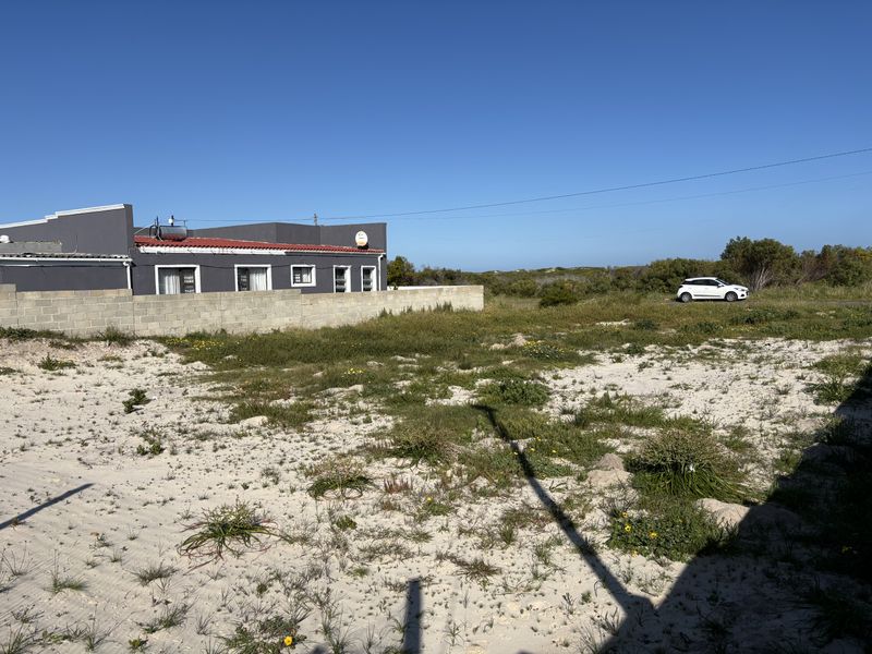 A view of a vacant residential plot featuring sandy terrain, natural vegetation, and a concrete block boundary wall adjacent to an existing structure.
