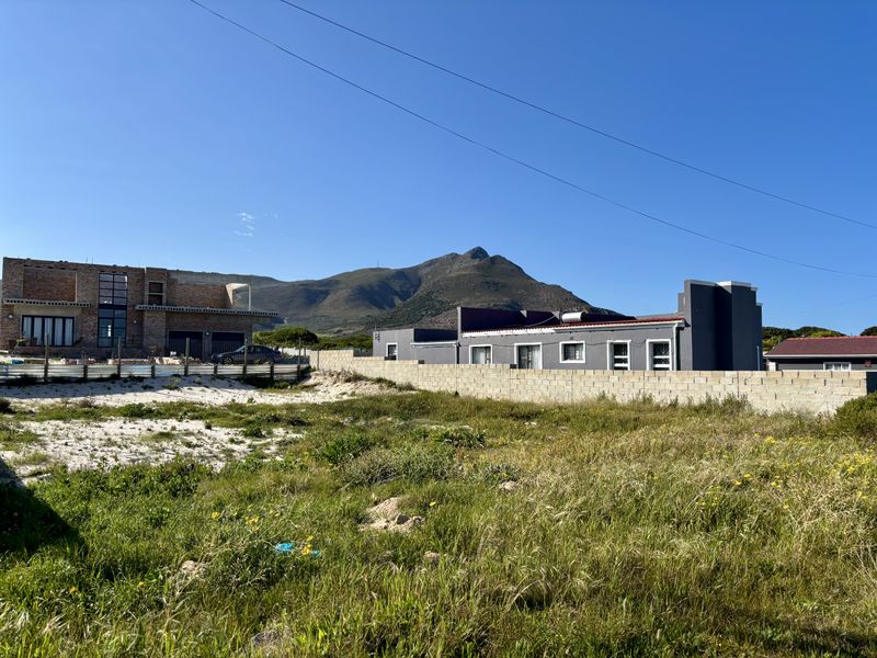 A vacant residential plot featuring natural vegetation and a boundary wall, with surrounding residential properties and mountain views in the background.