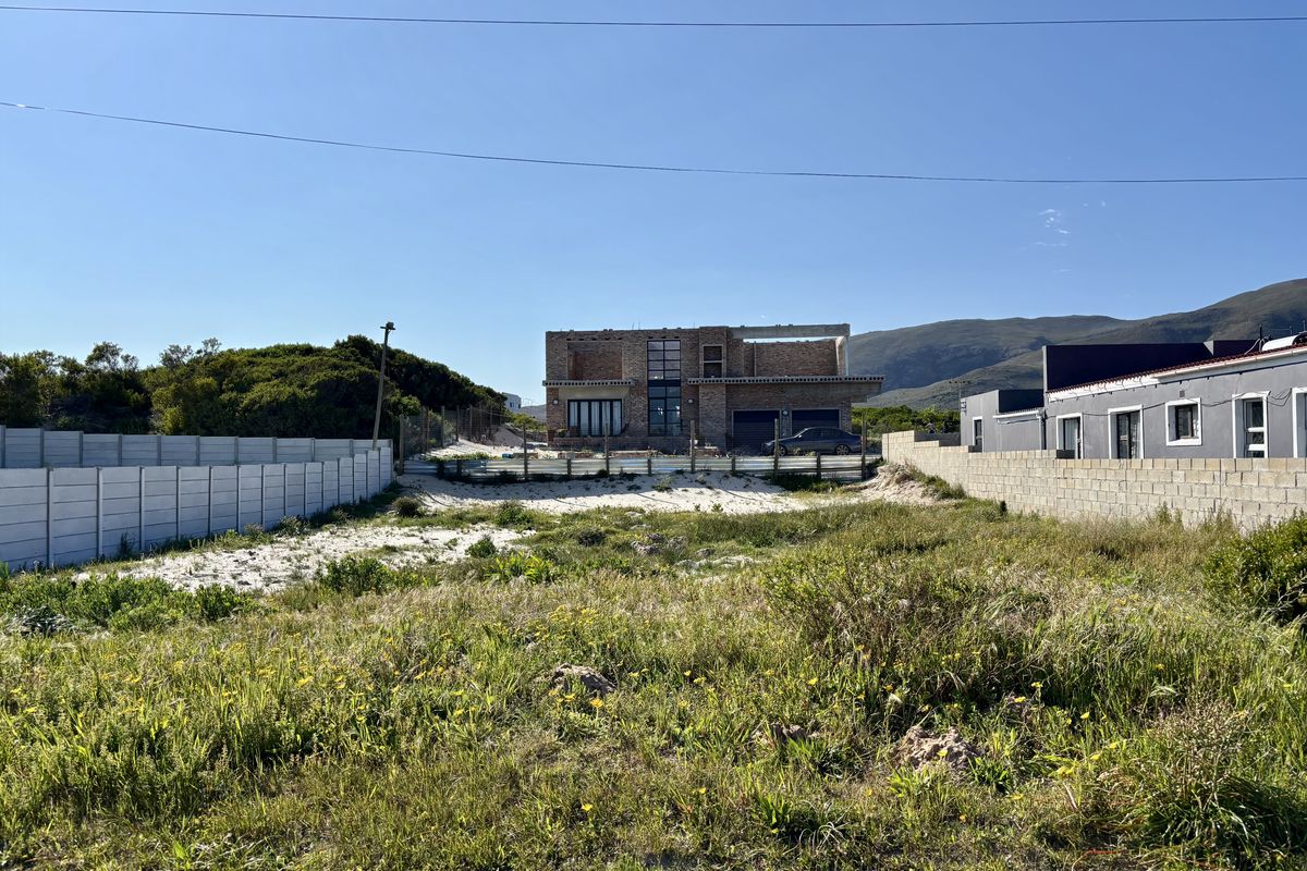 A vacant residential plot featuring sandy ground and precast concrete boundary walls, with a partially constructed building visible in the background.