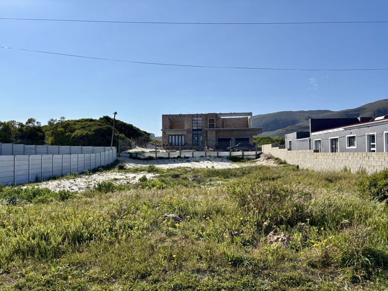 A vacant residential plot featuring sandy ground and precast concrete boundary walls, with a partially constructed building visible in the background.