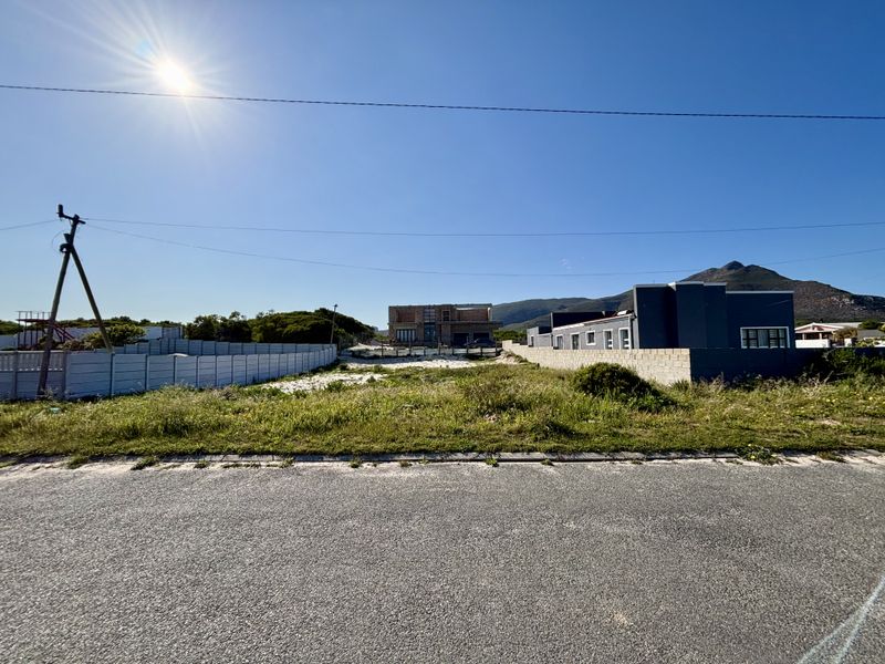 A vacant residential plot featuring a precast concrete boundary wall, situated along an asphalt road with mountain views in the background.