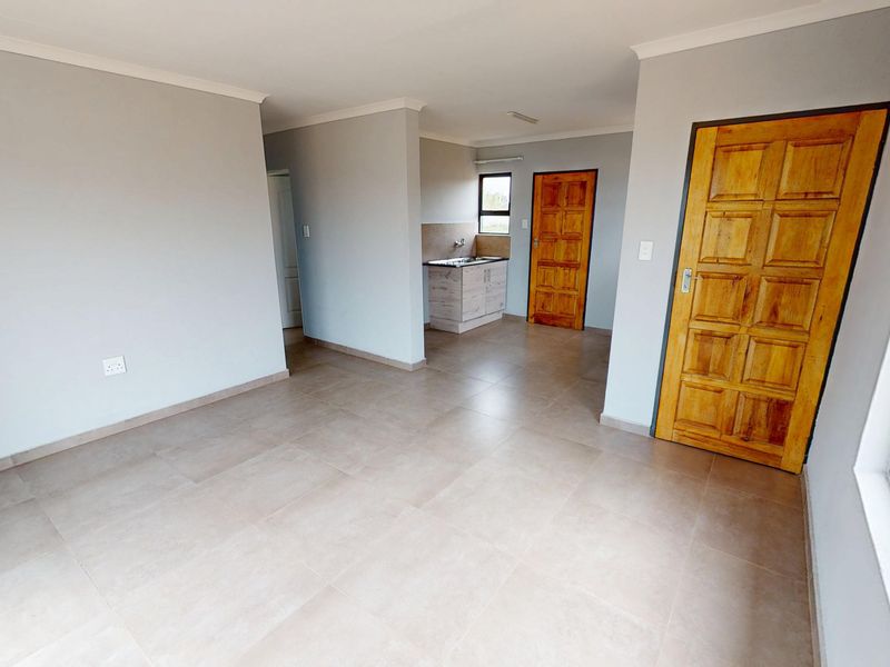 An open-plan living area featuring neutral tiled flooring, light grey walls, and a compact kitchen corner with built-in cabinetry and sink.